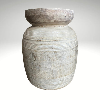 A rustic, light-colored wooden vase with a textured, ribbed surface and a natural wood rim sitting on a dark surface.