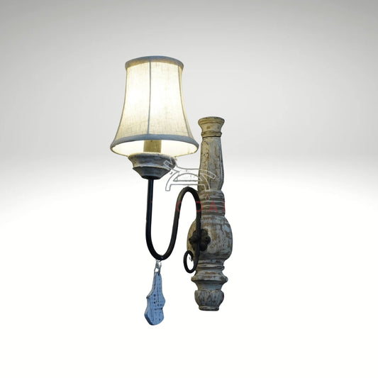 A distressed white wooden wall sconce with a curved black metal arm, a lighted cream fabric shade, and a small hanging wooden drop charm.