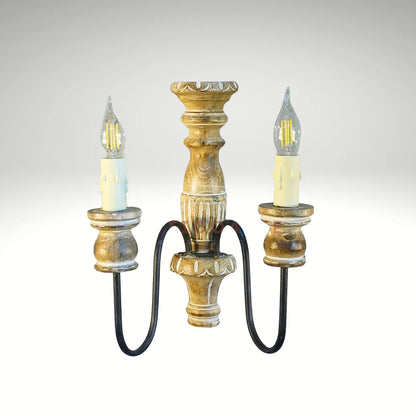 A vintage-style double wall sconce with a distressed carved wooden body, curved black metal arms, and two faux-candle LED bulbs.