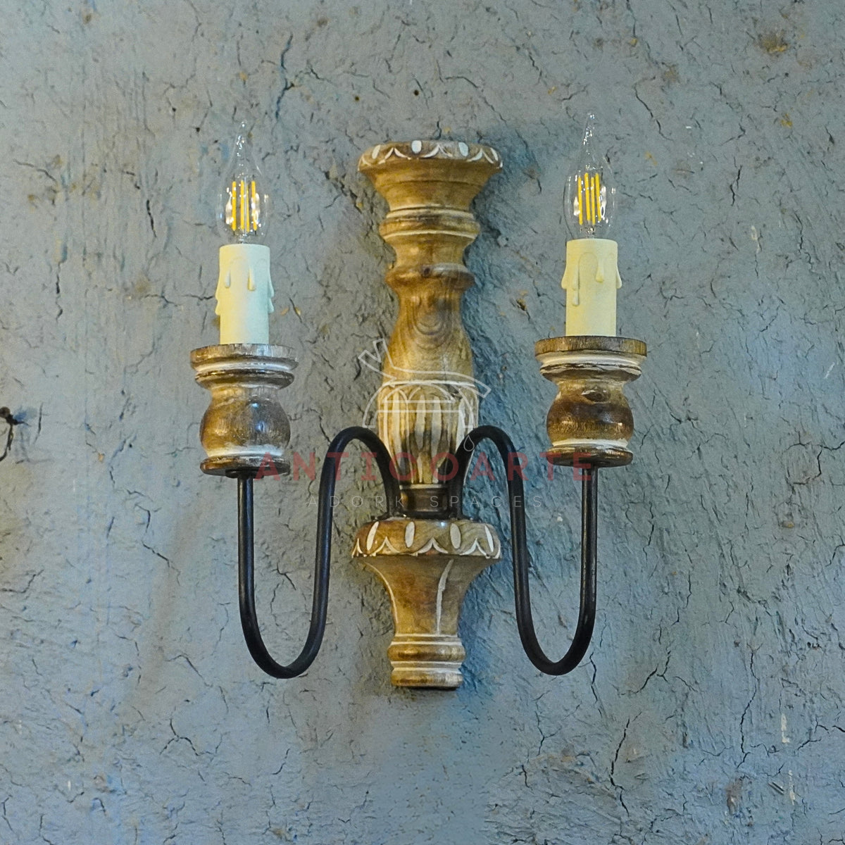 A vintage-style double wall sconce with a distressed carved wooden body, curved black metal arms, and two faux-candle LED bulbs.