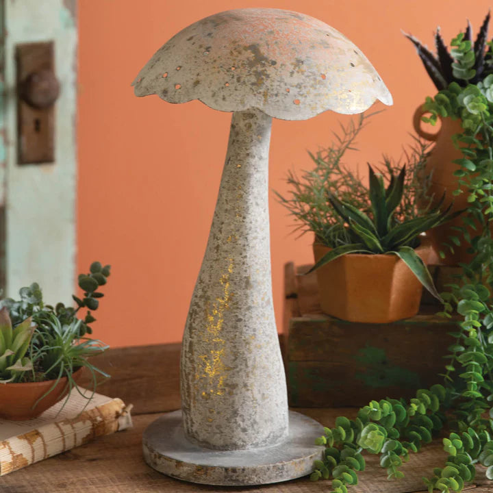 A rustic, mushroom-shaped decorative piece with a textured light-colored finish and golden highlights, placed on a wooden table alongside potted
