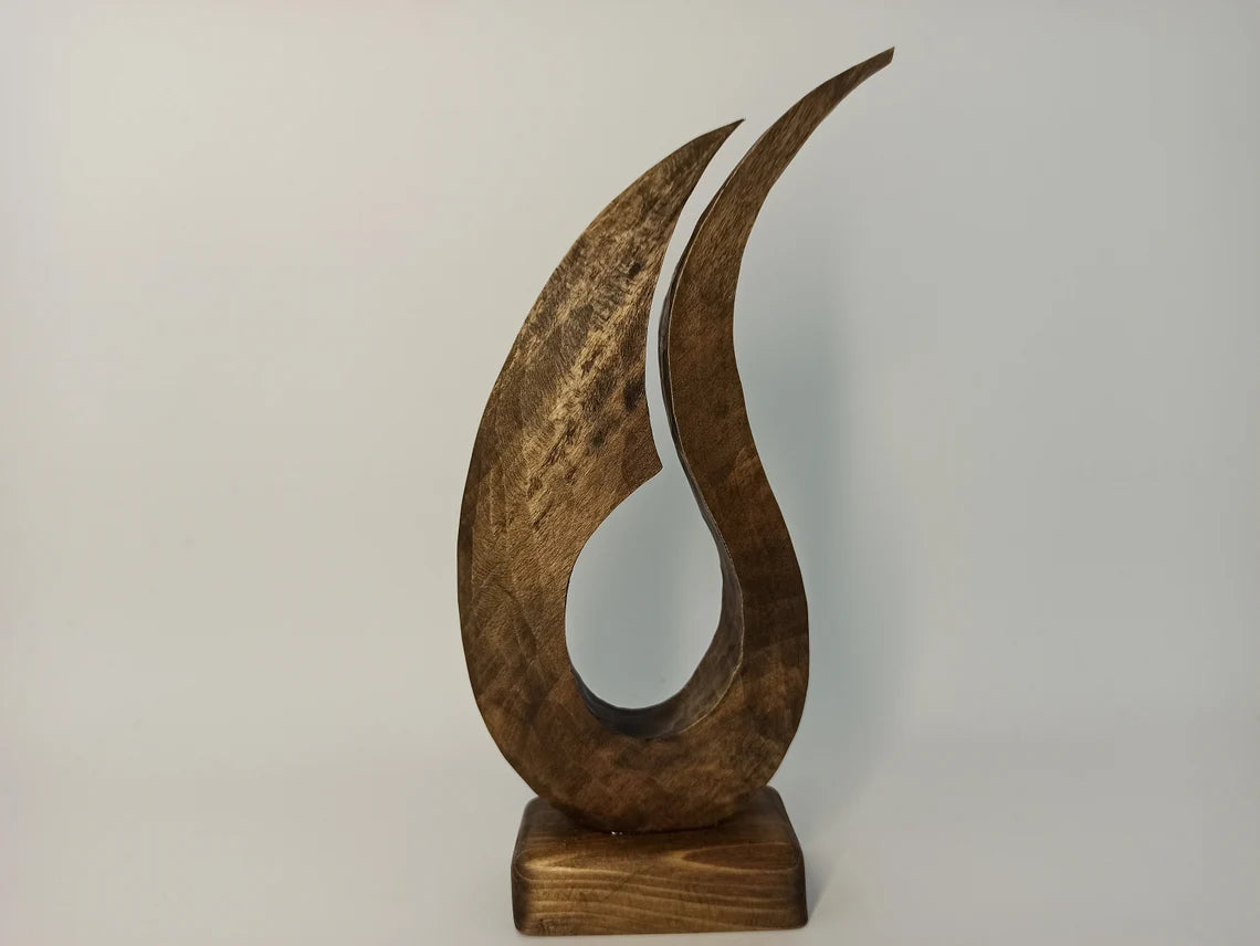 A contemporary abstract wooden sculpture shaped like a stylized, flowing flame with two tapering upward curves, mounted on a solid rectangular wooden base.
