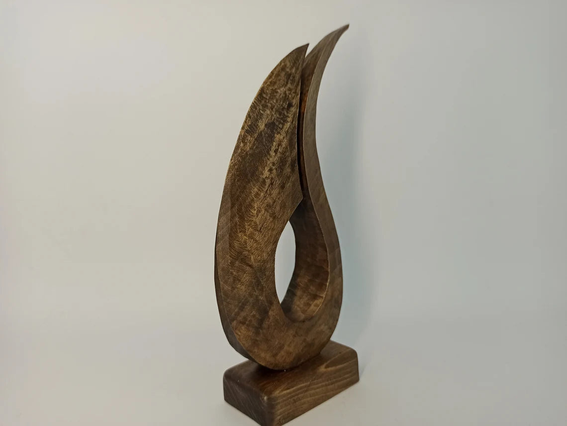 A contemporary abstract wooden sculpture shaped like a stylized, flowing flame with two tapering upward curves, mounted on a solid rectangular wooden base