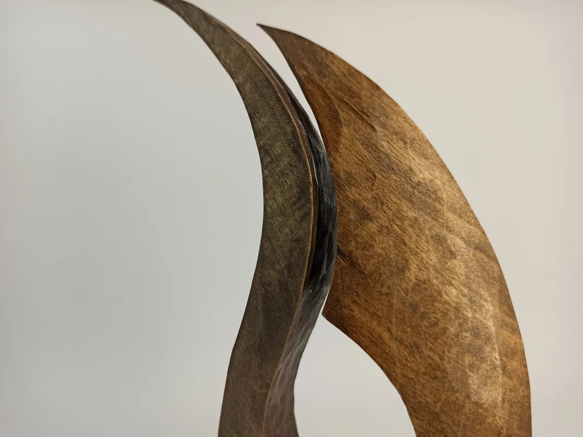 A contemporary abstract wooden sculpture shaped like a stylized, flowing flame with two tapering upward curves, mounted on a solid rectangular wooden base.