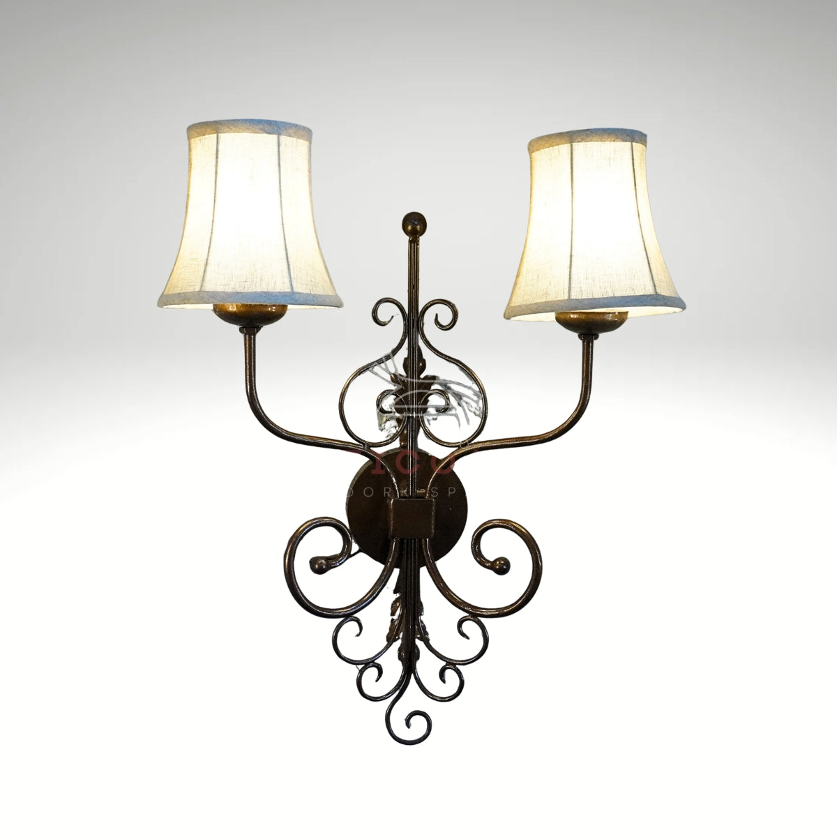 A rustic double-light wall sconce with a decorative dark metal scrollwork frame and two glowing cream fabric lampshades.
