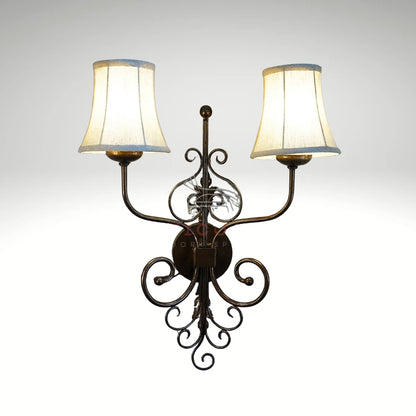 A rustic double-light wall sconce with a decorative dark metal scrollwork frame and two glowing cream fabric lampshades.