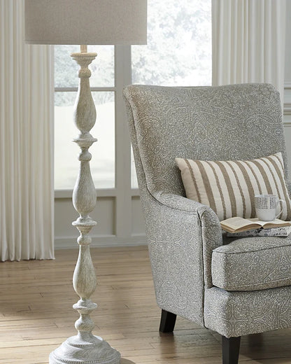 A distressed antique white floor lamp with a carved wooden-style base and a neutral beige fabric drum shade.