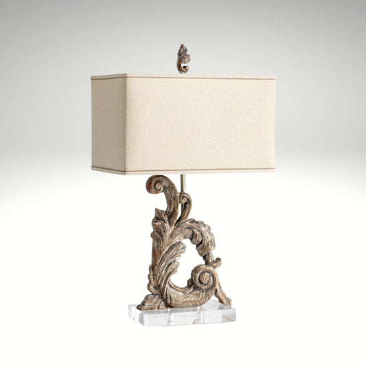 A table lamp featuring a carved acanthus leaf scroll base in an antique wood finish, a clear acrylic stand, and a rectangular linen shade.