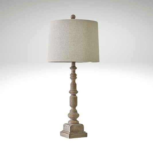 A traditional table lamp featuring a distressed, turned-wood candlestick base and a neutral beige fabric drum shade